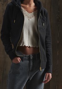 Felpa nera con zip e dettagli dorati, con testo ricamato. Indossata sopra un crop top crema con texture, bordo in pizzo e orlo sfrangiato. Jeans grigi a vita alta.