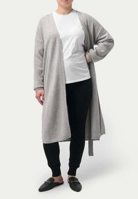 Grå lång cardigan i mjukt stickat tyg, med öppen framsida och ribbade ärmslut och fåll, bärs över en vit skjorta och svarta byxor.