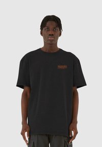 Sort bomulds T-shirt med rund hals, korte ærmer, oversized pasform og lille orange tekst på venstre bryst. Enkel design og glat tekstur.