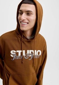Hnědá mikina s kapucí a stahovací šňůrkou, která má výrazný bílý a světle modrý nápis "STUDIO Jack & Jones." Materiál vypadá měkce a má přední kapsu.