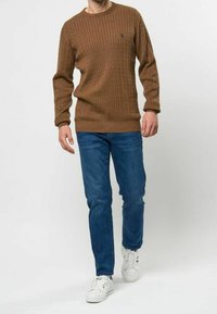 Pull en maille torsadée marron avec poignets et ourlet côtelés, porté avec un jean denim bleu et des baskets blanches. Le pull comporte un petit logo sur la poitrine.