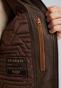 Giacca di pelle marrone con design trapuntato, caratterizzata da una cerniera arancione e un'etichetta con la scritta "AIR BRIDGE". La superficie è liscia con dettagli testurizzati.