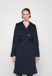 Trench coat bleumarin cu talie ajustată prin curea, design dublu-butonat, rever larg și nasturi negri; fabricat dintr-un material neted, rezistent la apă.