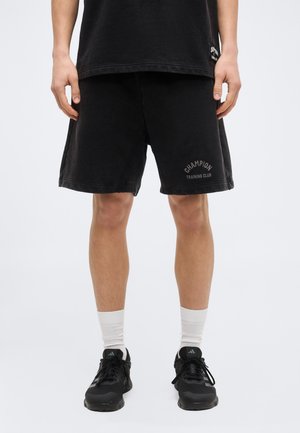 Άτομο που φοράει μαύρα σορτς Champion Training Club, μαύρα αθλητικά παπούτσια Adidas, λευκές κάλτσες και μαύρο T-shirt, στέκεται σε απλό υπόβαθρο.