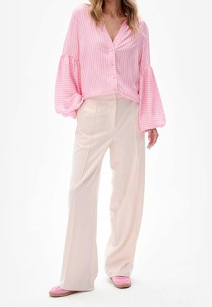 Femme portant un chemisier rose à rayures avec des manches volumineuses, un pantalon large crème et des chaussures plates roses sur fond blanc.