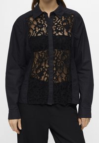 Chemise noire avec un panneau en dentelle transparente à l'avant, dotée d'un col classique et d'une patte de boutonnage. Manches longues et coupe structurée.