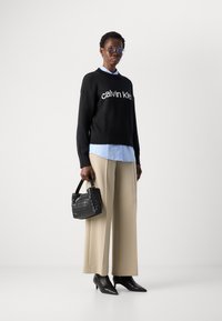 Person, der einen schwarzen Calvin Klein Pullover, ein hellblaues Hemd, beigen weit geschnittenen Hosen und schwarze spitze Stiefel trägt und eine schwarze geflochtene Handtasche hält.