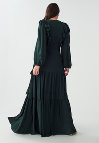Dark green, long-sleeve flowy dress featuring ruffled accents on shoulder and cuffs, tiered skirt, and a fitted bodice with a waist tie.