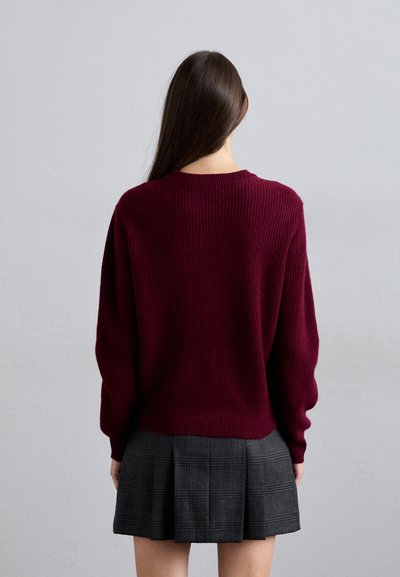 Donna con lunghi capelli castani che indossa un maglione bordeaux e una gonna plissettata grigia a quadri, in piedi di fronte a una parete grigia chiaro uniforme.