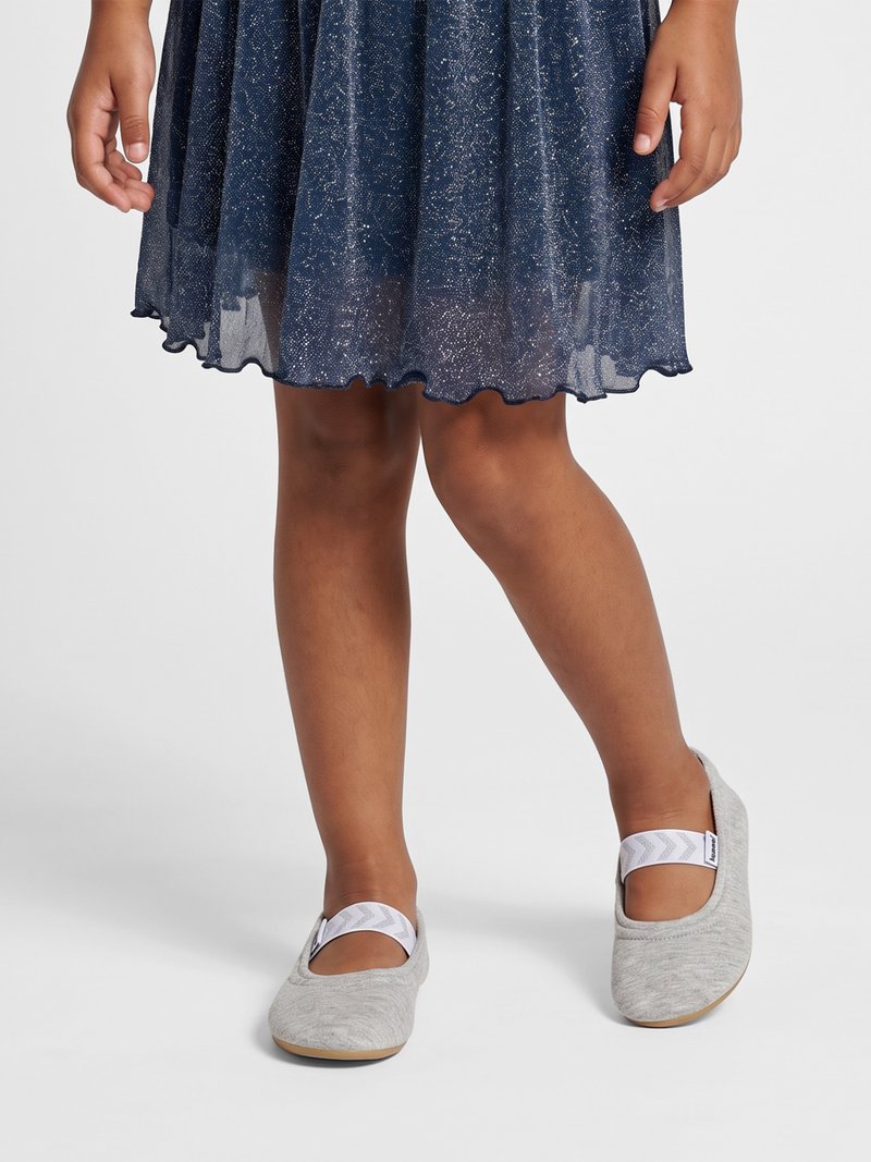 Kind trägt ein funkelndes dunkelblaues Kleid und hellgraue Mary Jane Schuhe mit weißen Riemen und steht auf einer weißen Fläche.
