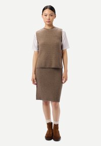 Femme aux cheveux foncés portant un gilet en tricot marron texturé et une jupe assortie, une chemise blanche à manches courtes, des chaussettes blanches et des bottines en daim marron.