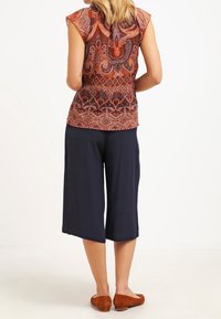 Top sin mangas impresa con un cálido patrón de paisley en rojo, naranja y marrón, combinado con culottes sueltos de color negro marino y planos marrones.