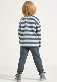 Bambino con capelli biondi di lunghezza media che indossa una maglietta a maniche lunghe grigia e azzurra a righe, pantaloni grigio scuro e scarpe alte grigie, in piedi.
