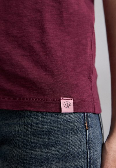 Burgunderfarbenes T-Shirt aus weichem Stoff, mit einem kleinen rosa Etikett am Saum, das ein geometrisches Logo zeigt, kombiniert mit einer dunklen Jeans.