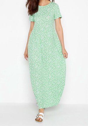 Kobieta nosząca jasnoróżową sukienkę maxi z krótkim rękawem w biały wzór kwiatowy i białe sandały z klamrą, stojąca na jednolitym białym tle.