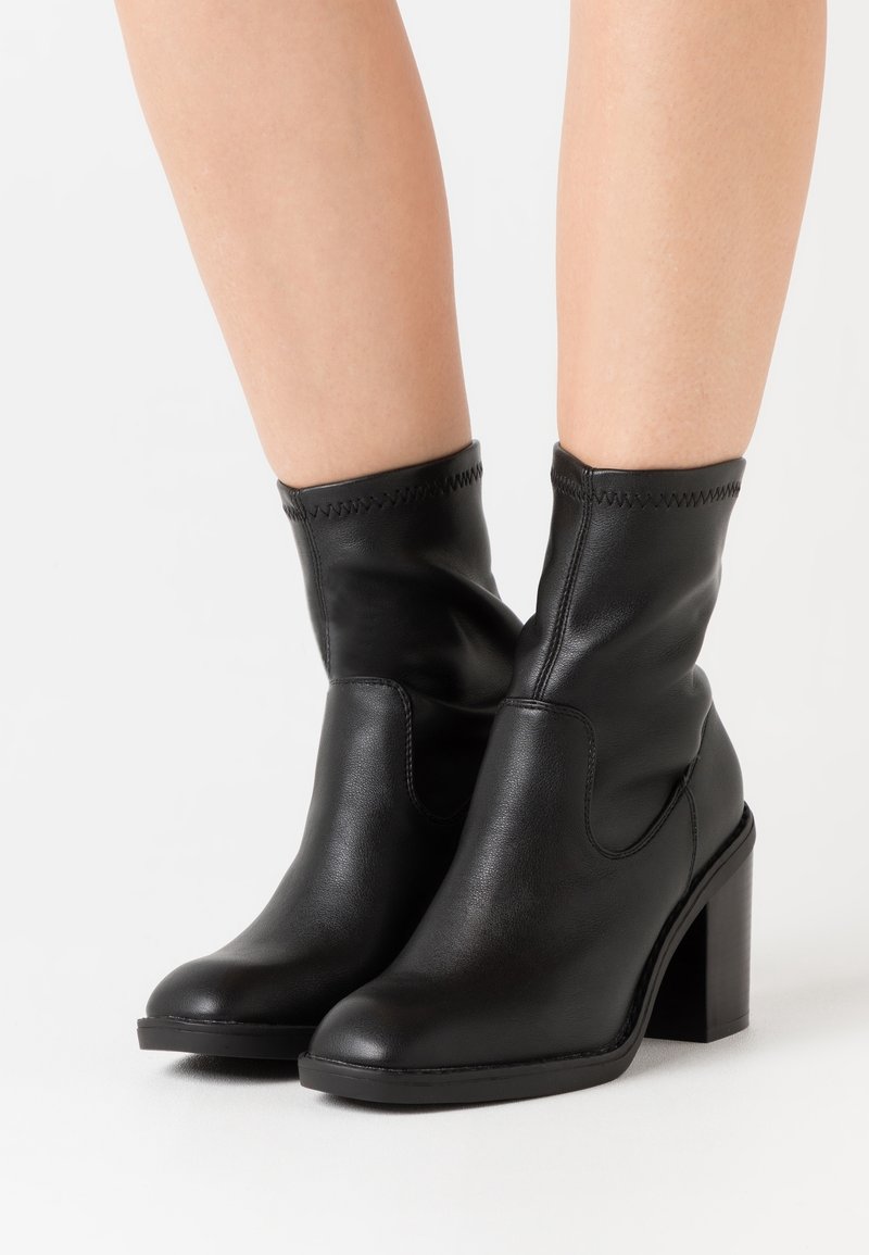 Bottes basses en cuir noir avec un bout arrondi, des côtés élastiques, des accents cousus et un talon bloc épais. Texture lisse et design élégant.