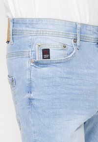 Jean en denim bleu clair porté par une personne, montrant la poche arrière avec l’étiquette de la marque Kordshi et une partie d’une chemise blanche au-dessus.