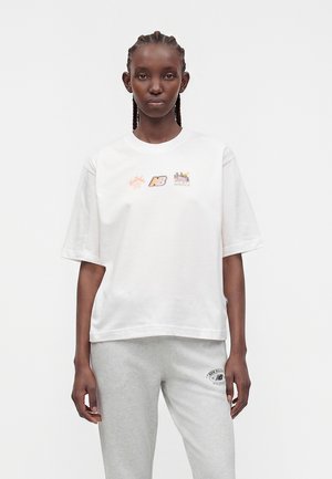 Γυναίκα με πλεξούδες, φορώντας λευκό t-shirt με πολύχρωμα λογότυπα και ανοιχτό γκρι φόρμα με μαύρο λογότυπο στον αριστερό μηρό.