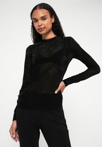 Top preto de mangas longas feito de um tecido transparente e texturizado. Apresenta uma silhueta ajustada e punhos canelados, combinado com parte inferior preta.