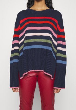 Mujer que lleva un suéter de punto azul marino con rayas horizontales multicolores y borde rosa, combinado con pantalones de cuero rojos.