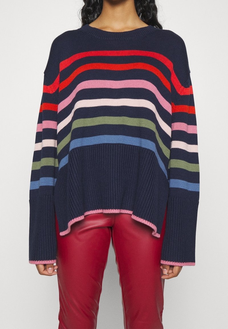 Femme portant un pull en tricot bleu marine avec des rayures horizontales multicolores et des bordures roses, associé à un pantalon en cuir rouge.