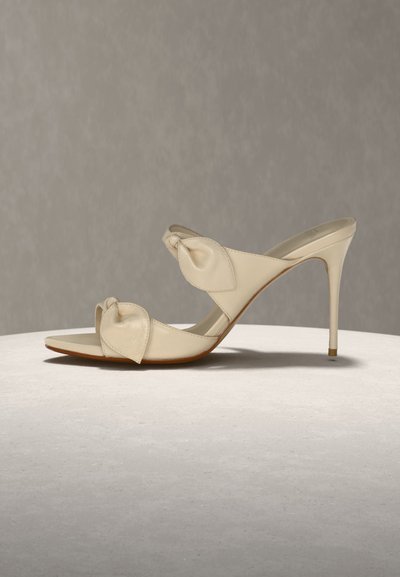 Μπεζ mule πέδιλο με ψηλό τακούνι στιλέτο, δύο λουριά με κόμπους, σε ουδέτερη επιφάνεια και φόντο.