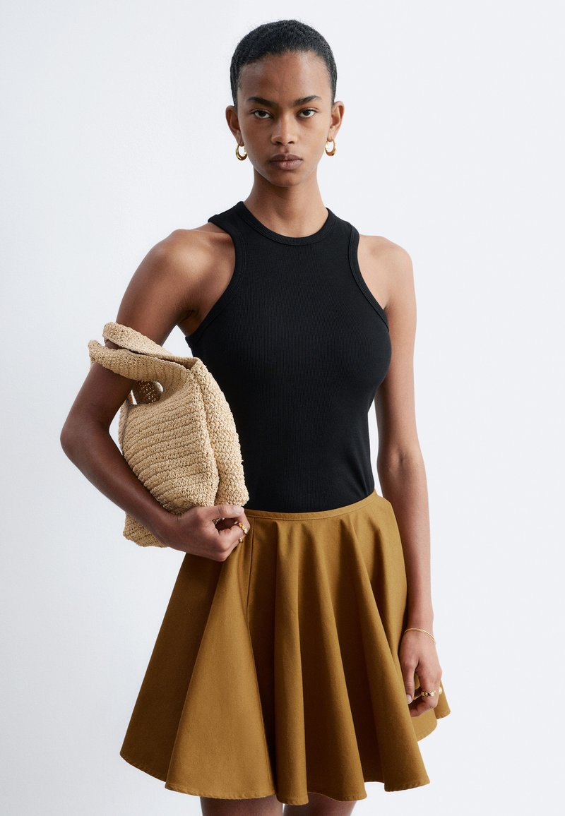 Femme en haut noir sans manches et jupe marron tenant un sac tressé beige sur un fond blanc uni.
