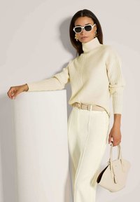 Vrouw in een crèmekleurige geribbelde turtleneck, witte broek, beige riem, met een beige handtas, draagt witte zonnebril en gouden oorbellen, leunend op een witte cilinder.