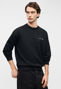 Μαύρο φούτερ με στρογγυλή λαιμόκοψη από βαμβακερό μείγμα ύφασμα, με μικρό λευκό λογότυπο στο στήθος και ριμπ μανσέτες και στρίφωμα.