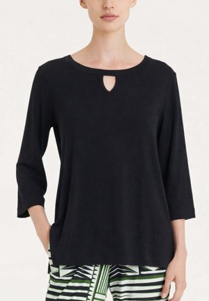 Donna con indosso un top nero a maniche lunghe con un piccolo taglio a goccia sul collo e pantaloni a fantasia neri, bianchi e verdi.