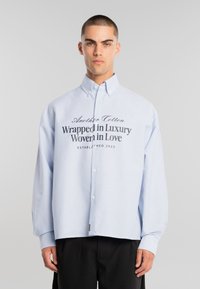 Joven con cabello corto que lleva una camisa de botones de rayas azul claro con el texto "Envuelto en lujo, tejido con amor" y pantalones negros.