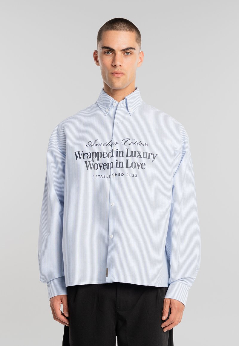 Joven con cabello corto que lleva una camisa de botones de rayas azul claro con el texto "Envuelto en lujo, tejido con amor" y pantalones negros.