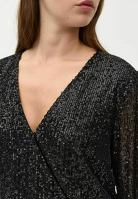 Fekete flitteres ruha mély V-kivágással, mely texturált felülettel és lebegő fazonnal rendelkezik, a fényt visszaverő flitterekkel ragyog.