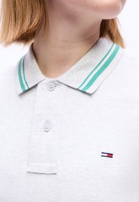 Άτομο που φορά ανοιχτόγριζο πόλο μπλουζάκι με γιακά ριγέ πράσινου χρώματος και μικρό ορθογώνιο λογότυπο κόκκινου, λευκού και μπλε στο στήθος, το κεφάλι γυρισμένο στο πλάι.