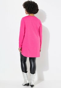 Tunique rose à manches longues avec un ourlet arrondi, associée à des leggings noirs brillants et des bottes de cowboy argentées, vue de dos.