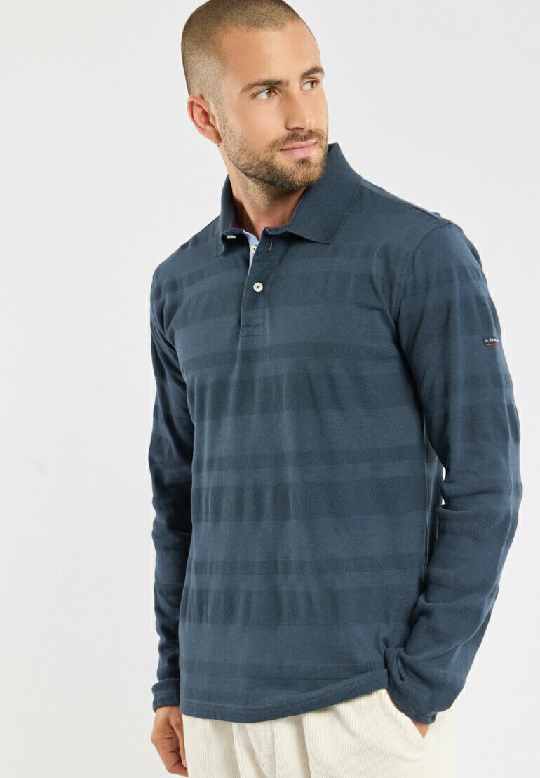 Homme avec des cheveux courts et une barbe portant un polo à manches longues bleu foncé et un pantalon clair, se tenant avec une main dans la poche.