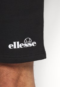 Shorts en tissu noir avec le logo blanc « ellesse » imprimé près de l'ourlet au-dessus d'une jambe poilue visible.