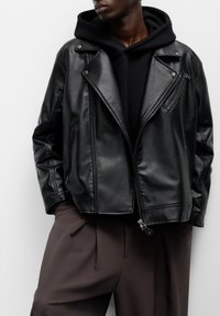 Crna kožna motoristička jakna s patentnim zatvaračem sprijeda i bočnim džepovima, nošena preko crne dukserice. Sadrži izrezani ovratnik i srebrne metalne detalje.