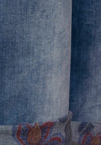 Detalní pohled na modrou denimu s ohrnutým manžetou, odhalující barevnou květinovou výšivku na vnitřním okraji.