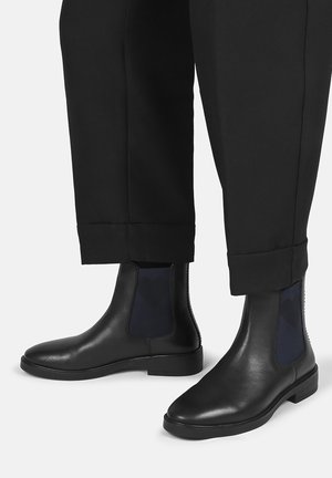 Black leather Chelsea boots with dark elastic side panels worn with black cuffed trousers on a light background.