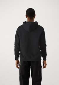 Black hoodie with a drawstring, textured fabric, and a kangaroo pocket. Worn with dark denim pants, seen from the back.