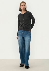 Donkergrijze gebreide cardigan met knopen aan de voorzijde, gecombineerd met lichtblauwe wijde jeans. Zwarte schoenen met grote strikaccenten.