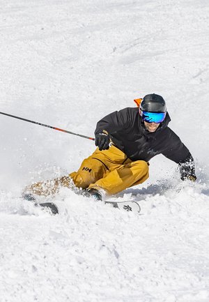 Skiër in een donkere jas en gele broek, sneeuw spuit omhoog. Voorzien van een zwarte helm en spiegeltieten, op een met sneeuw bedekte helling.