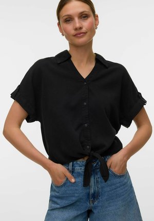 Frau mit hellem Teint und zurückgebundenem Haar, trägt ein kurzärmeliges schwarzes Hemd mit Knöpfen, das an der Taille gebunden ist, und blaue Jeans, Hände in den Taschen.