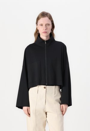 Veste cropped noire avec un col haut, fermeture éclair à l'avant et deux grandes poches à rabat. Le tissu semble lisse avec une coupe décontractée. Portée sur un pantalon beige.