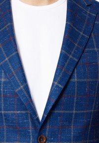 Niebieska marynarka w kratę z teksturowanej tkaniny. Posiada brązowy guzik, pod spodem biała koszulka z okrągłym dekoltem oraz kontrastujące czerwone i żółte linie.