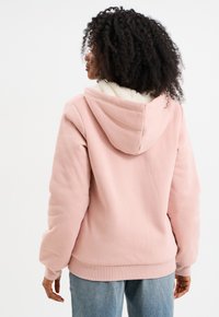 Felpa rosa chiaro con un morbido rivestimento testurizzato, dotata di cappuccio e polsini a coste. Indossata con jeans di denim blu, vista da dietro.
