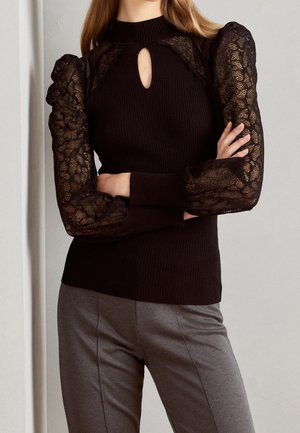 Un pull en laine côtelé noir avec un col roulé, doté de longues manches en dentelle bouffantes et d'une découpe en forme de clé au niveau du col. Porté avec un pantalon gris.
