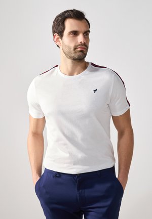 Weißes Baumwoll-T-Shirt mit kurzen Ärmeln und navyblauem, gesticktem Logo. Mit roten und navyblauen Streifen auf den Schultern, runder Halsausschnitt.