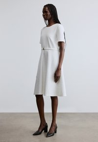 Robe blanche texturée avec manches courtes, taille ceinturée et jupe évasée. Associée à des escarpins noirs à bout pointu. Design simple et élégant.